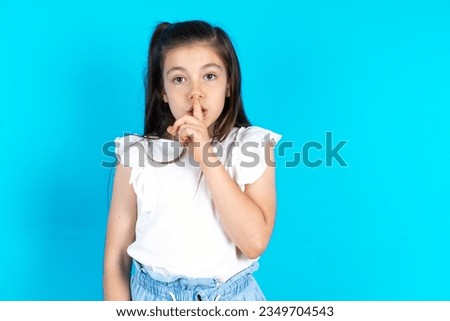 Surprised caucasian kid girl wearing white t-shirt makes silence gesture, keeps finger over lips and looks mysterious at camera