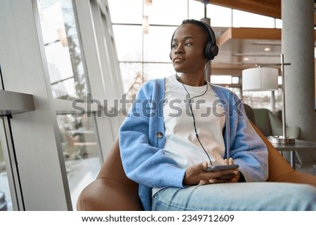 African Black teenage girl university or college student refugee sitting in chair in university campus, coworking office space or airport lounge, wearing headphones looking away at window. Royalty-Free Stock Photo #2349712609