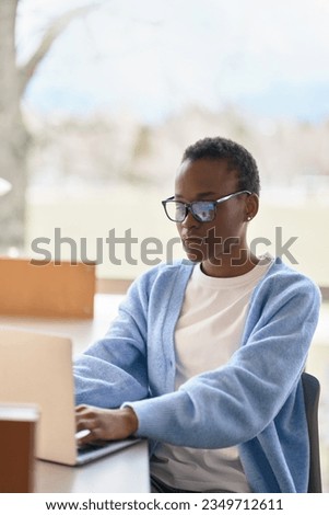 Busy African Black girl student wearing eyeglasses using laptop computer technology sitting at desk. Serious young woman elearning or working looking at pc, watching webinar, typing on pc, vertical.
