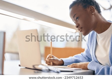 African Black girl student using laptop computer in university campus sitting at desk hybrid learning online, doing college course study or research, watching virtual class, taking notes, elearning.