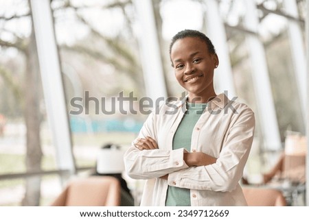Smiling short-haired confident ethnic Black girl college student standing indoors. Portrait. Happy cute African schoolgirl with arms crossed looking at camera in modern foreign university campus. Royalty-Free Stock Photo #2349712669