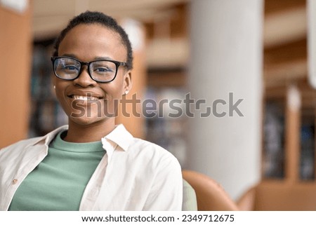 Happy cute African teen girl, smiling short-haired cute Black ethnic college student wearing eyeglasses looking at camera in modern university campus library. Close up face portrait. Royalty-Free Stock Photo #2349712675