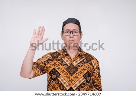 A southeast asian man pledges loyalty and makes an oath. Wearing a batik shirt and songkok skull cap with hand on chest. A middle aged muslim male in his 40s. Isolated on a white background. Royalty-Free Stock Photo #2349714609