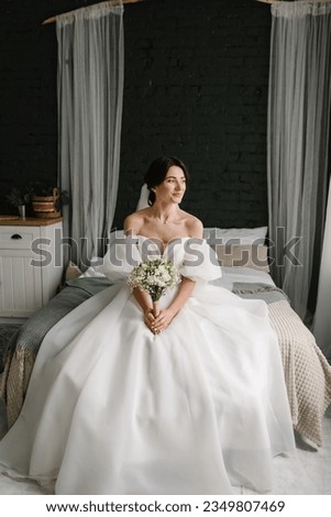 Young fashion model with perfect skin and professional makeup. A bride in stylish dress sitting in bed at home. Luxury wedding day. Fashion photo. Waiting for the groom. Trendy wedding style shot.