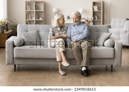 Happy older couple of grandparents enjoying leisure, conversation on home sofa together, talking, laughing, discussing retirement, elderly healthcare, family news, having fun. Full length shot