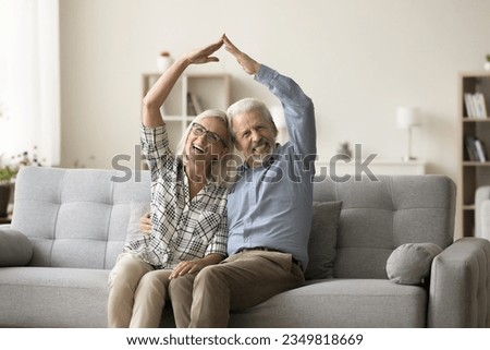 Happy older retired couple making roof hands overhead, looking at camera, sitting on home sofa, hugging, smiling for portrait, showing symbol of housing, protection, safety, insurance