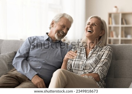Cheerful elder husband and wife having fun at home together, relaxing on cozy couch at home, talking, laughing, enjoying retirement, conversation, family leisure, humor