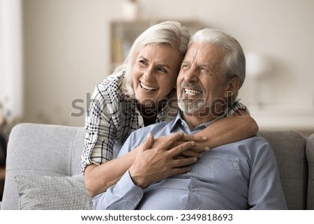 Dreamy elderly aged couple in love hugging at home, laughing, looking away in happy thoughts, thinking on good retirement plans, discussing vacation, enjoying marriage, relationships Royalty-Free Stock Photo #2349818693