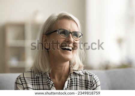 Happy beautiful blonde mature woman in elegant glasses sitting on couch, looking away, smiling, laughing with open mouth, showing healthy white teeth. Happy elderly lady enjoying leisure at home Royalty-Free Stock Photo #2349818759