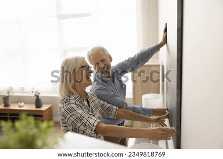 Joyful positive retired husband and wife enjoying interior decoration work, hanging large artwork on wall, holding picture together, smiling, laughing, having in new apartment