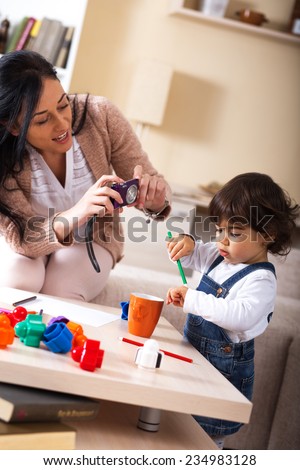 Mother taking a picture of her baby girl.They playing with toys in living room.