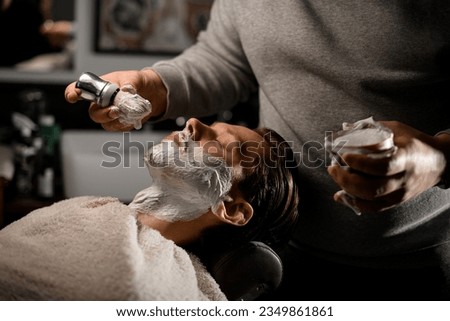 Hairdresser applying shaving foam on client's beard before shaving in barber shop. Side view. Man's service Royalty-Free Stock Photo #2349861861