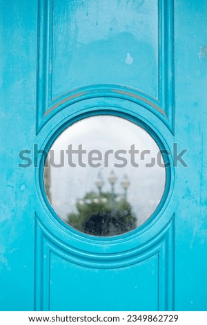 Turquoise blue bright painted wooden door with circle round window into music concert venue reflection in glass of green tree lamppost with three lights and vague silhouette of person photographer