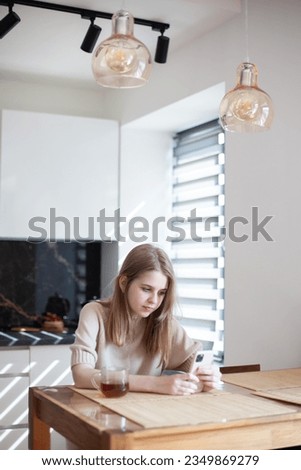Teen girl sitting at table with cup of tea using smartphone at home. Remote education, online courses for teens, online shopping, good morning concept. Neutral colors. Vertical image.