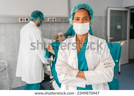 Photo of black beautiful midwife wearing surgical cap, mask and gloves standing with arms crossed while preparing for childbirth in operating theatre. Royalty-Free Stock Photo #2349870447