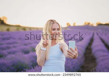 Lavender field. Pretty woman with blond hair walking between rows of lavender flowers with mobile phone, taking selfie, communicating in social networks, relaxing, resting, enjoying nature
