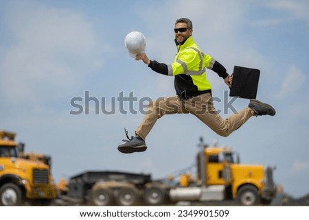 Middle aged builder excited jump on site construction. Excited builder construction worker in a safety helmet jumping in front of the trucks. Excited crazy builder man in helmet jump outdoor. Royalty-Free Stock Photo #2349901509