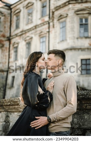 Woman in black long dress kiss man stand on stairs near ancient palace at sunset. Female, male walk near brick walls in street. Luxury couple newlyweds near old Castle. Stylish bride hug groom outdoor