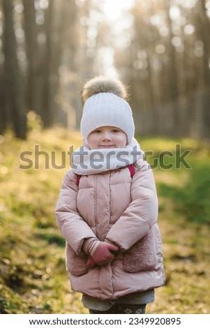 Child stands in background of golden bokeh sun in forest. Happy girl walking at sunset in autumn park. Children on street near trees. Autumn photo, start winter. Kid in warm clothes walking outdoors.