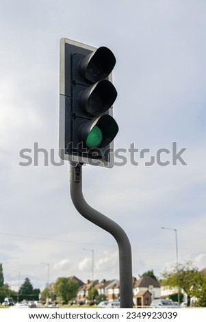 Traffic light signal green bent curving pole post in residential street outside of London, England on a dry sunny mildly cloudy day with houses, cars, lampposts, trees and shrubs in the background