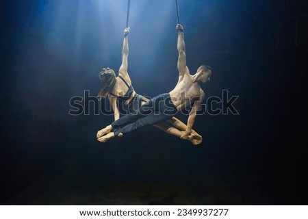 Aerial straps duo performance: a man and woman execute graceful acrobatic feats in mid-air against a black background, dressed in black and illuminated by a white-blue glow Royalty-Free Stock Photo #2349937277