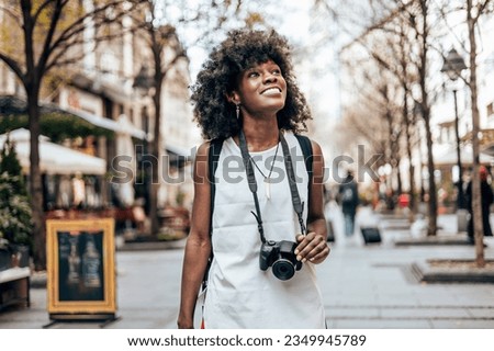 Young black female tourist enjoys walking through the streets of a beautiful European city. She is happy and using her photo camera to take fantastic architecture photographs. Bright sunny day.