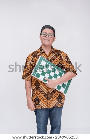 An indonesian man holding a full-sized chessboard. An amateur or professional chess player at a tournament. Wearing a batik shirt and songkok skull cap. Isolated on a white background.
