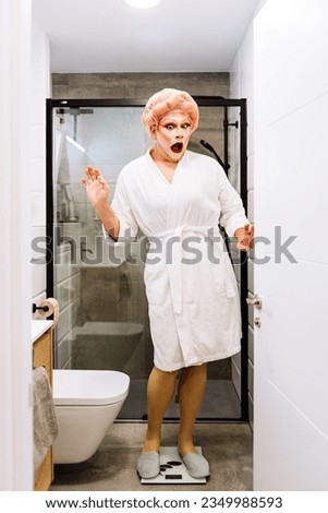 Full body of shocked transgender female with bright makeup in white bathrobe standing on scales in bathroom Royalty-Free Stock Photo #2349988593