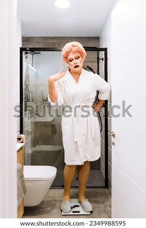 Full body of worried transgender female with bright makeup in white bathrobe standing on scales in bathroom Royalty-Free Stock Photo #2349988595