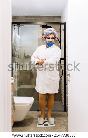 Full length of Drag Queen with crossed arms wearing white bathrobe and standing on scales in bathroom Royalty-Free Stock Photo #2349988597