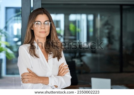 Confident 45 year old Latin professional mid aged business woman corporate leader, mature female executive manager wearing eyeglasses standing in office arms crossed looking at camera, portrait.