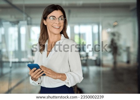 Happy smiling mature middle aged professional business woman executive or entrepreneur wearing eyeglasses holding smartphone using cell phone standing in office lobby, looking away at copy space.