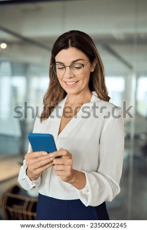 Smiling middle aged business woman holding cell phone standing in office. Happy mature professional businesswoman wearing eyeglasses holding smartphone using smartphone looking at cellphone, vertical.