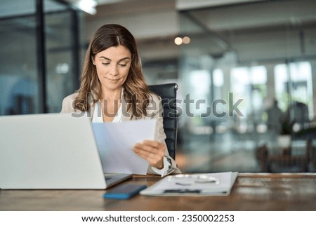 Busy mid aged business woman working in office with laptop reading document. Mature professional female manager lawyer attorney holding paper finance report sitting at desk in office. Copy space Royalty-Free Stock Photo #2350002253