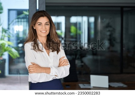 Smiling confident Latin professional mid aged business woman corporate manager leader, happy beautiful 40s mature female executive standing at office window arms crossed looking at camera, portrait.