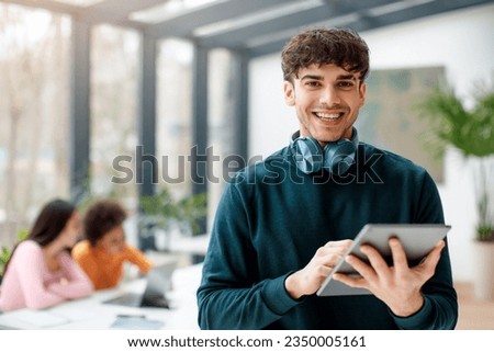 Portrait of happy european student guy holding digital tablet for video lesson or educational app, studying with classmates in classroom, ladies sitting at table on background Royalty-Free Stock Photo #2350005161