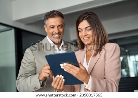 Smiling busy middle aged business man and business woman professional corporate executive leaders wearing suits discussing digital strategy using tablet computer standing in office working together.