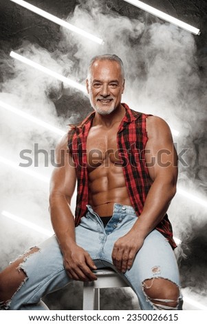 Muscular middle-aged male model with a trendy grey beard, shirtless, wearing an unbuttoned checkered shirt and ripped jeans, sits on a black cube, with lights and smoke in the background