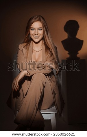 Stunning and captivating fashion model in an elegant beige suit, posing gracefully on a chair against a backdrop of warm lighting and a silhouette of an ancient Greek bust