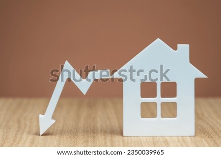 Reducing the interest rate for a mortgage or lowering the cost of insurance for real estate. White house model and graphic arrow pointing down close-up on a brown background. copy space. Royalty-Free Stock Photo #2350039965