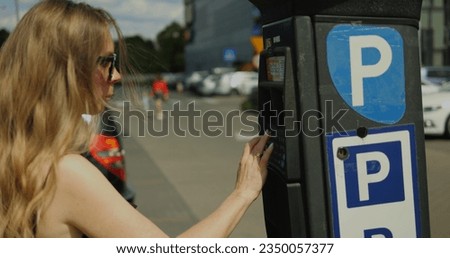 Stylish lady in sunglasses enter license plate number on parking meter outdoors. Beautiful female adult student in sundress with long brown hair pay for car parking at parking machine.