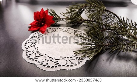 Black frame, white round napkin, green pine branch, red flower on background with reflection and sparkle of light. Abstract location for photo shoot. Cchristmas table decor. Concept of winter holiday