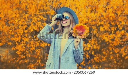 Autumn color style outfit, stylish young woman photographer with film camera and yellow maple leaves wearing round hat and coat in the park