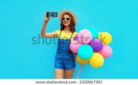 Summer image of happy laughing young woman taking selfie with mobile phone holds bunch of colorful balloons having fun wearing straw hat on blue studio background