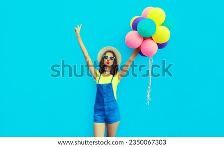 Summer image of happy cheerful young woman with bunch of colorful balloons having fun wearing straw hat on blue studio background