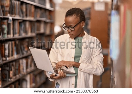 Busy African Black girl student wearing eyeglasses using laptop standing in university or college library among bookshelves looking at computer searching information online. Authentic shot