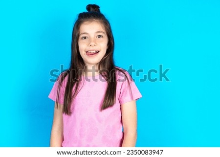Funny caucasian kid girl wearing pink T-shirt over blue background makes grimace and crosses eyes plays fool has fun alone sticks out tongue.