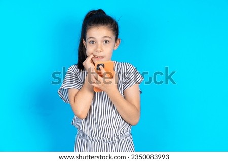 Afraid funny caucasian kid girl wearing striped dress over blue background holding telephone and bitting nails