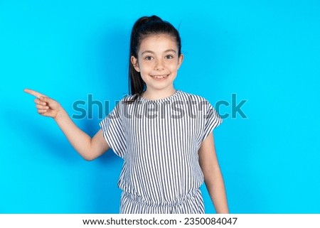 Positive caucasian kid girl wearing striped dress over blue background with satisfied expression indicates at upper right corner shows good offer suggests to click on link