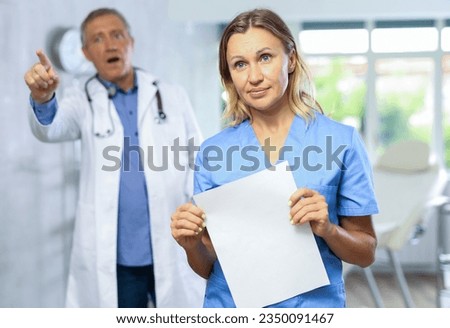 Middle-aged nurse standing with sad look in cabinet while angry mature doctor is scolding her behind Royalty-Free Stock Photo #2350091467